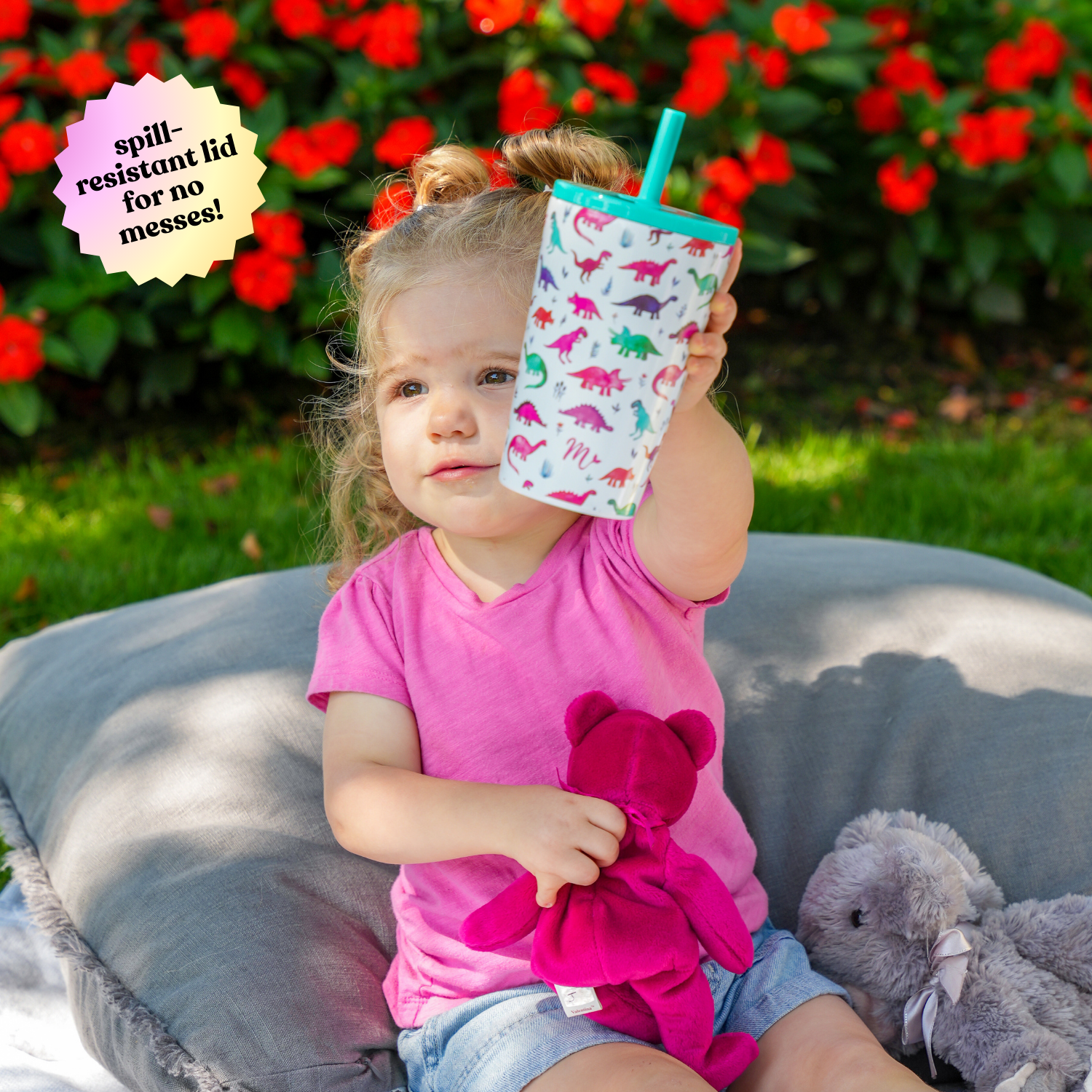 Young girl sitting outdoors on a cushion, holding up a 14 oz stainless steel cup with a colorful dinosaur design, featuring a spill-resistant lid, with vibrant red flowers in the background.