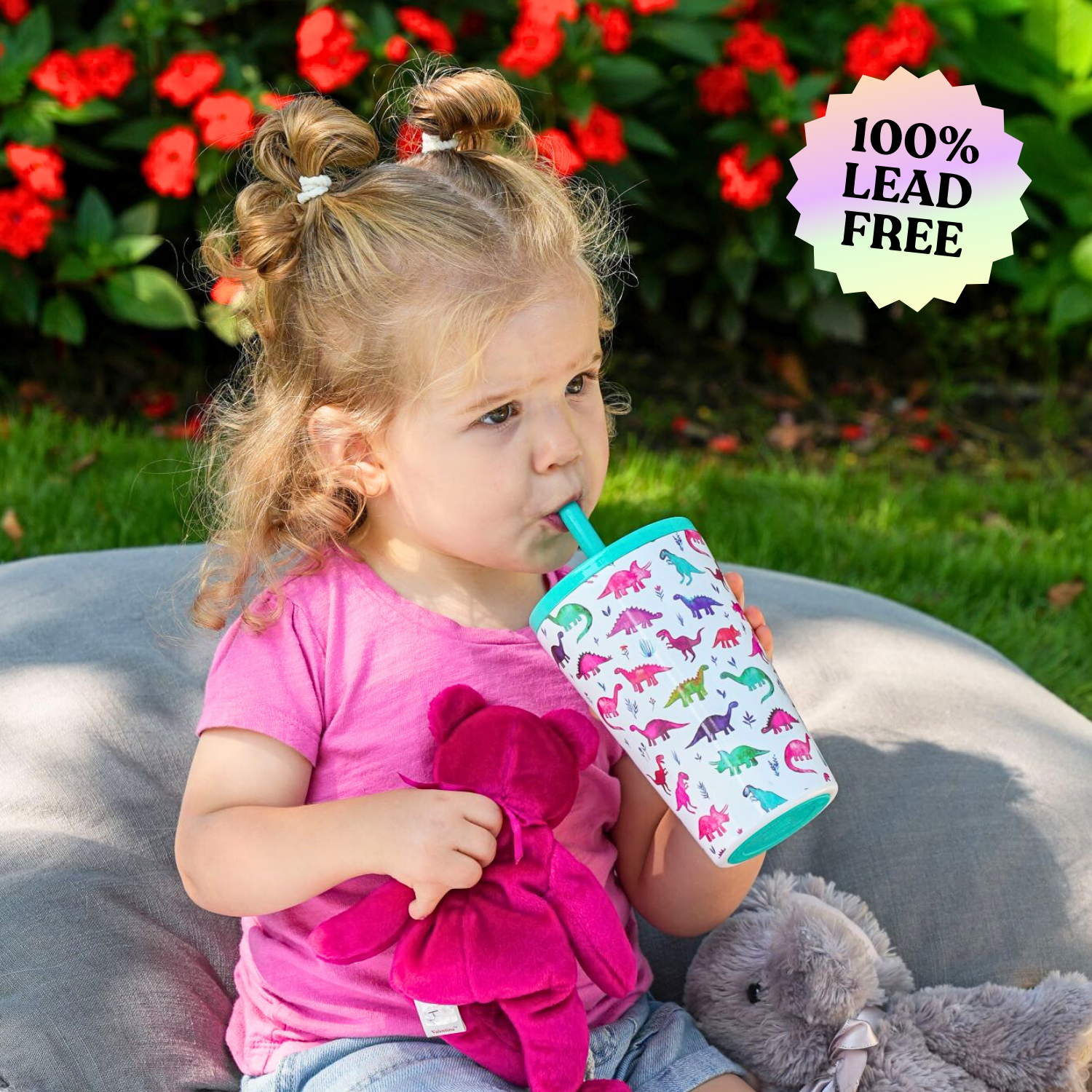 Young girl drinking from 14oz tiny dinos stainless steel kids cup while sitting on a pillow in a park holding a stuffed animal.