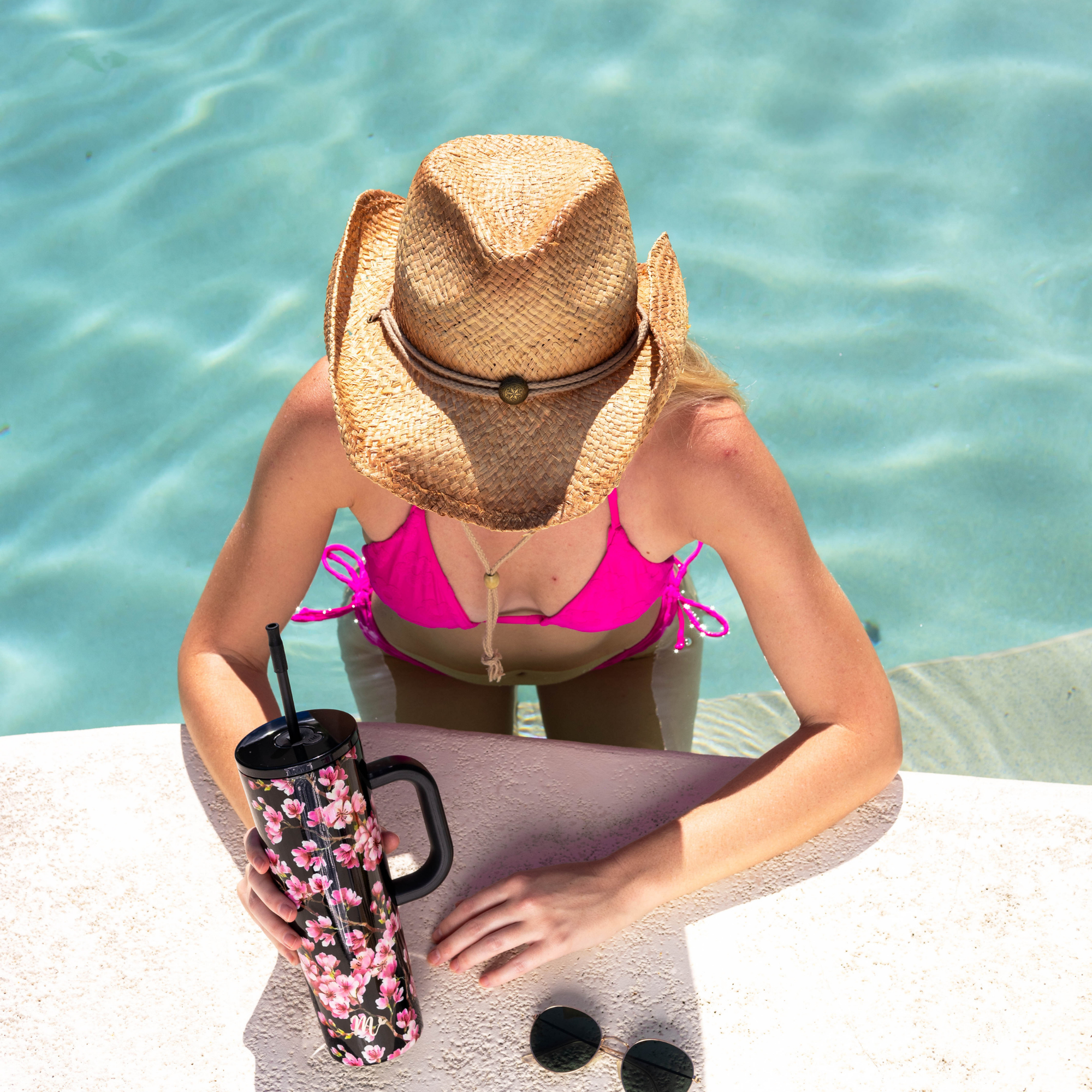 Top view of a person in a pink bikini and straw hat relaxing in a pool, holding a black 30oz tumbler with a pink cherry blossom design and black handle. The person is leaning on the pool edge, with sunglasses placed next to the tumbler.
