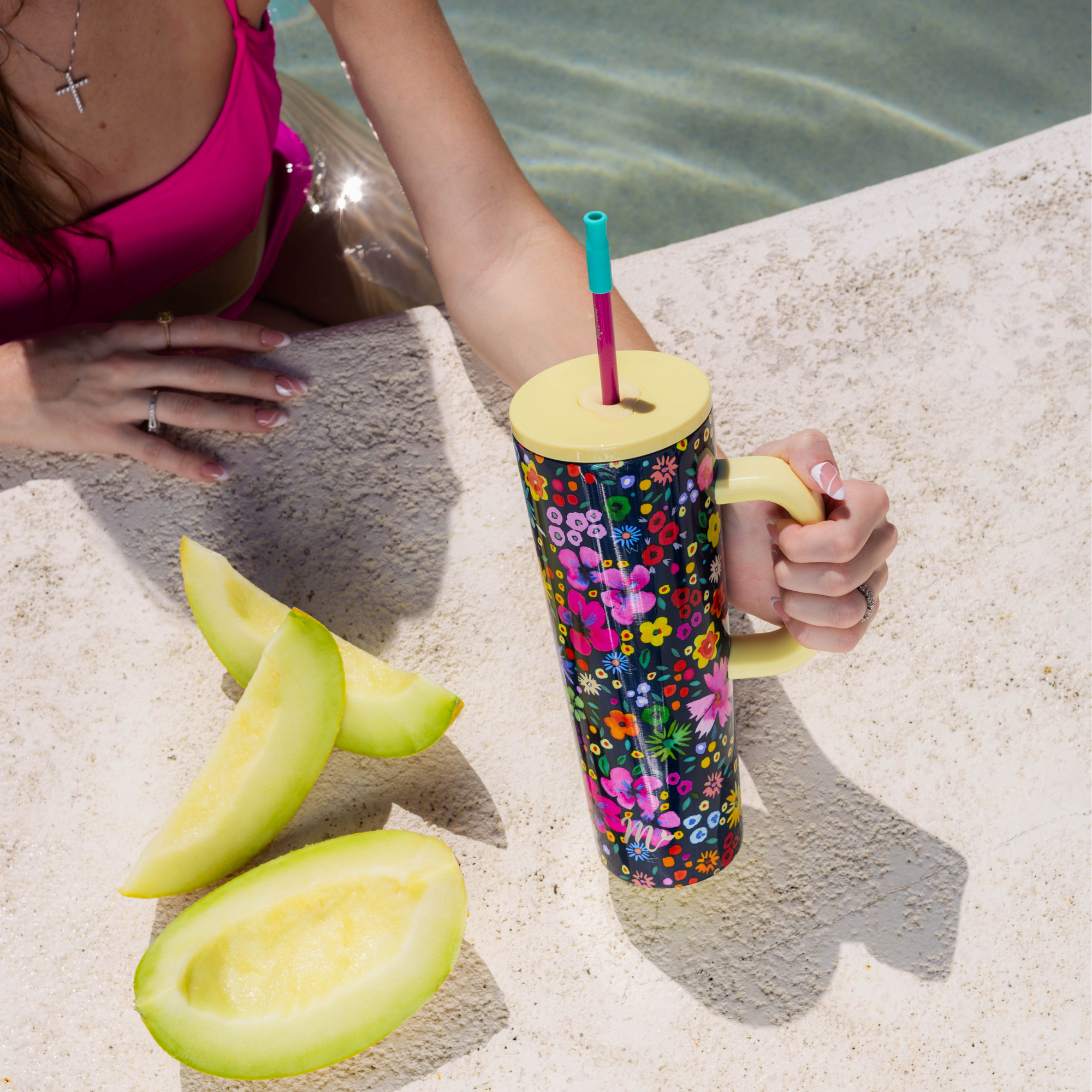 Woman in a pink bikini enjoying poolside with slices of melon, holding a 30oz tumbler with a vibrant ditzy floral design, yellow handle, and pink straw with a teal silicone tip. The image highlights the tumbler's bold style, perfect for refreshing moments by the water.
