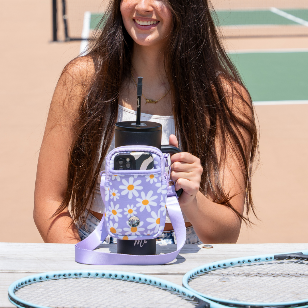 30 oz Tumbler with Purple Daisy Sling at the Tennis Court - A smiling woman enjoys a break at the tennis court, holding a black tumbler with a matching purple daisy-patterned sling. The sling provides storage space for essentials like a phone, making it perfect for staying hydrated and organized during sports and outdoor activities.