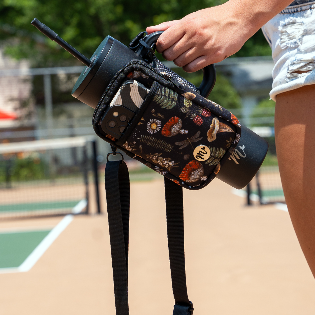 30 oz Black Tumbler with Nature-Inspired Sling - A close-up of a person carrying the tumbler using the neoprene sling, which features a woodland design with mushrooms, ferns, and berries. The pouch provides extra storage, shown holding a smartphone, and the adjustable strap offers hands-free convenience for active lifestyles.