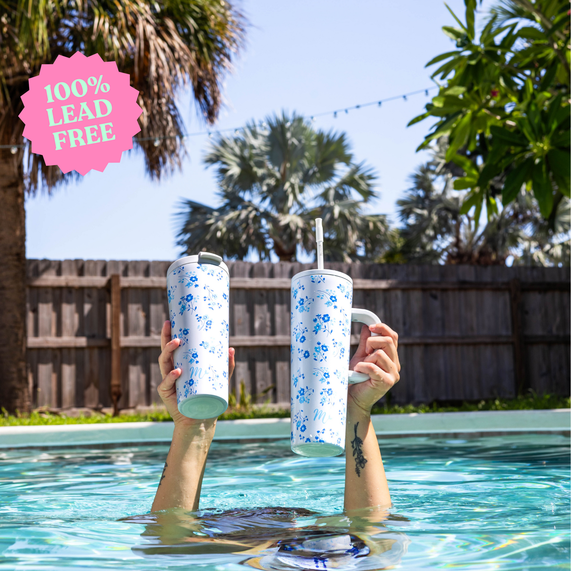 Two hands holding up 30oz tumblers with blue forget-me-not floral designs in a pool setting, highlighting one with a handle and straw and the other with a fliptop lid. The image showcases the tumblers' 100% lead-free feature and their stylish, practical design for outdoor and poolside use.