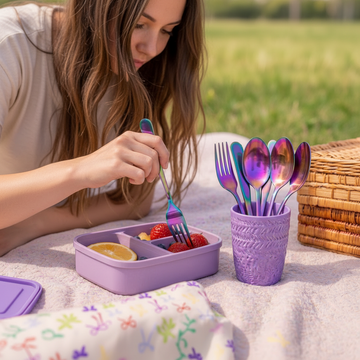 Person holding a colorful fork over a purple bento box with food, next to a container of more utensils on a picnic blanket.