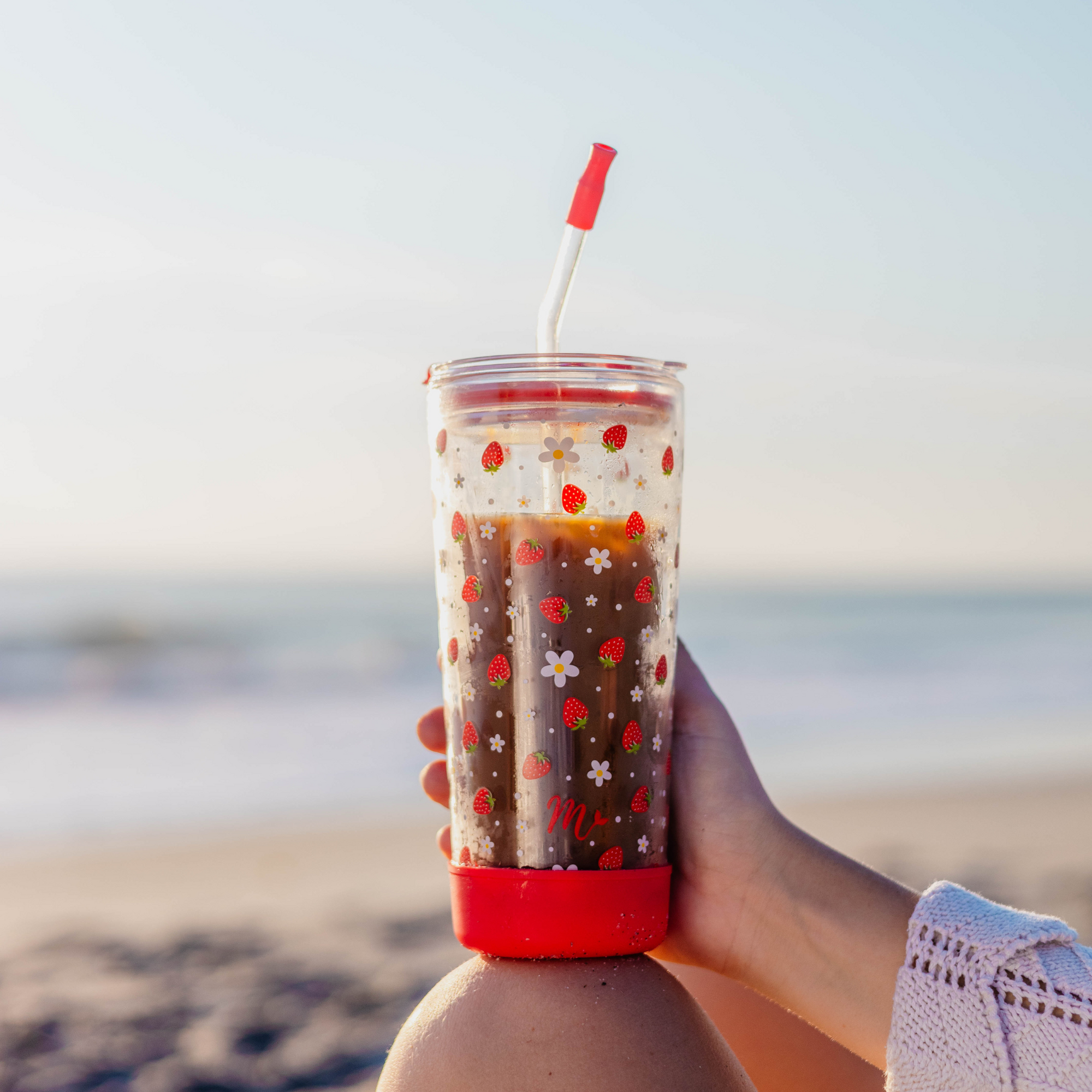 Person holding a 20oz double-wall glass cup with a strawberry design, filled with iced coffee, on a beach.