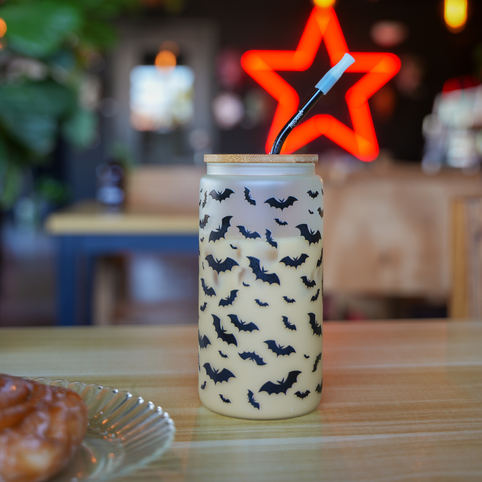 Frosted glass tumbler with black bat patterns, featuring a wooden lid and reusable straw, placed on a table with a neon star light in the background.