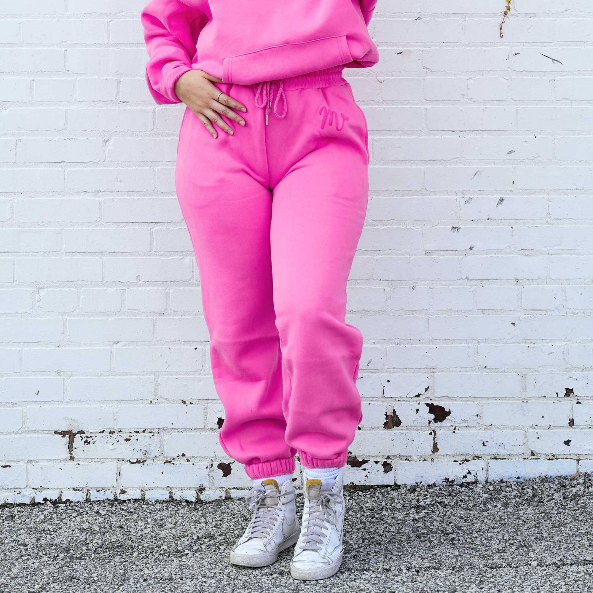 Model wearing bright pink sweatpants with an embossed logo on the left thigh, paired with white high-top sneakers. The sweatpants have a comfortable fit with an elastic waistband and cuffs, showcasing a casual yet stylish look against a white brick wall