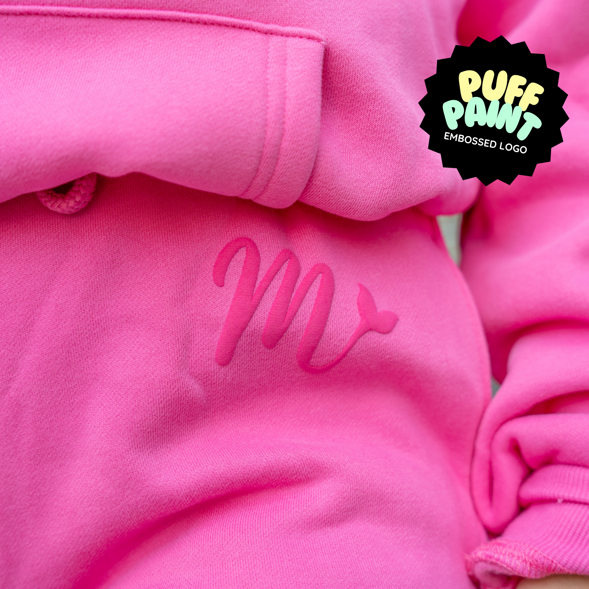 Close-up of the bright pink hoodie featuring a puff paint embossed logo. The design showcases a raised 'M' with a tail detail, highlighting the textured, vibrant logo that adds a unique touch to the hoodie