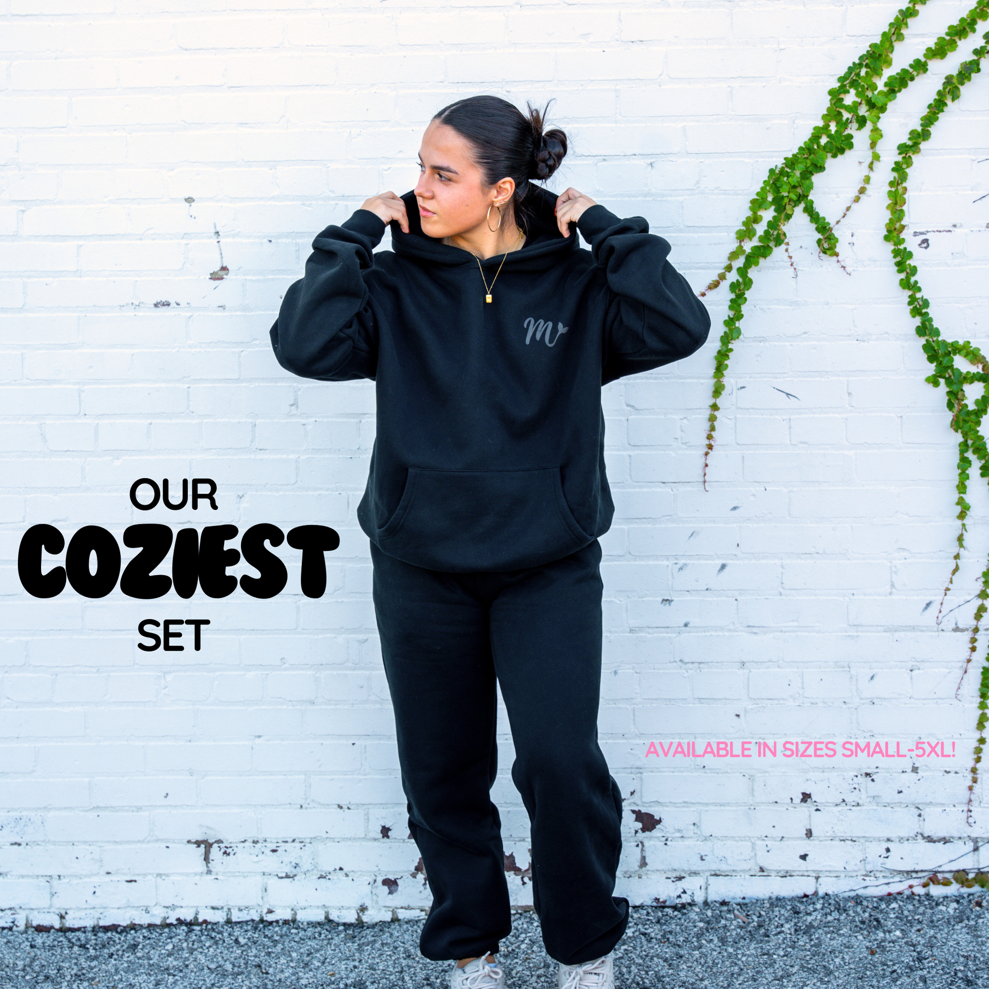 "Model wearing a black cozy hoodie and sweatpants set, both featuring a subtle embossed logo. The set is highlighted as 'Our Coziest Set,' available in sizes Small to 5XL. The model stands against a white brick wall with green vines, showcasing the comfortable and stylish outfit.