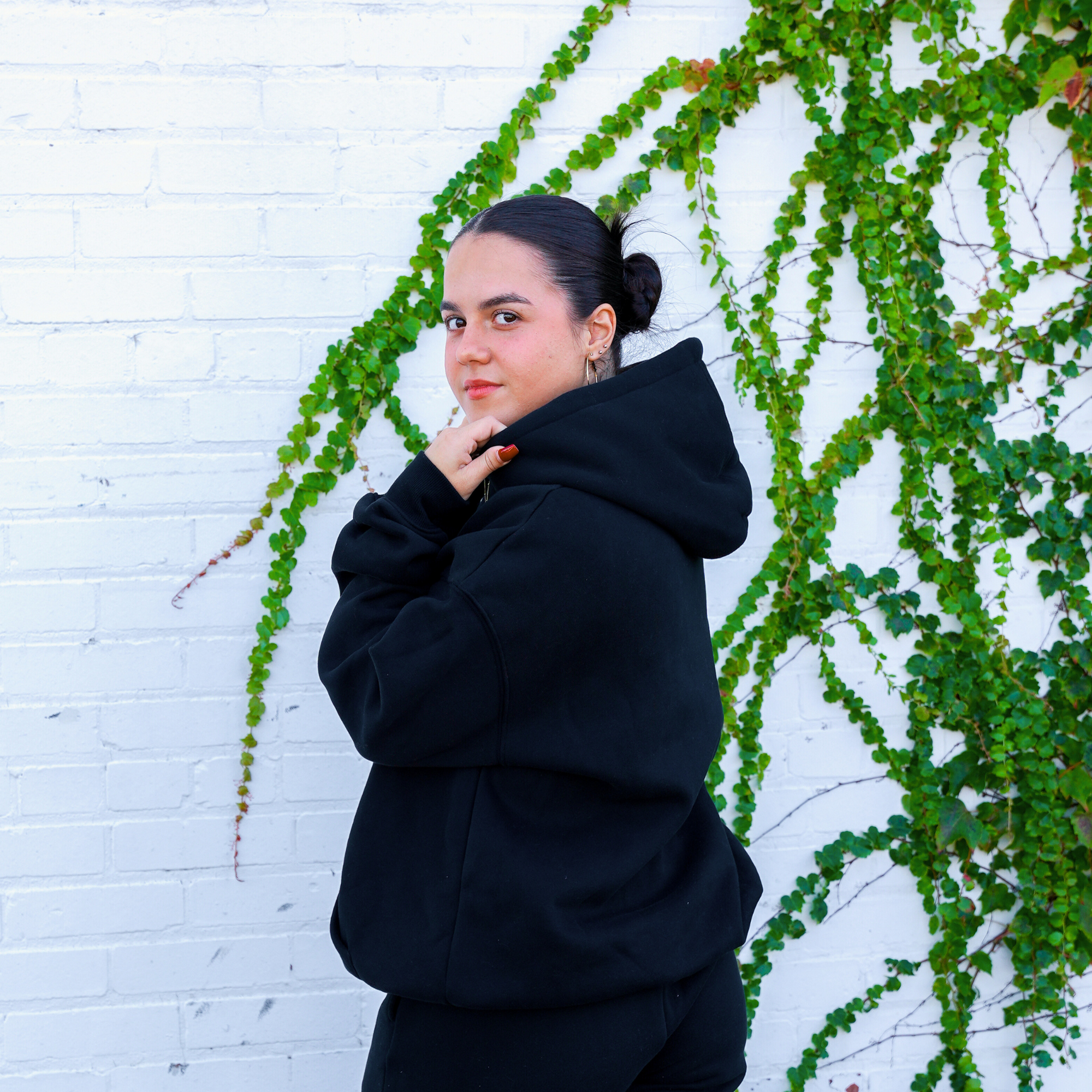 Model wearing a black hoodie, standing in profile against a white brick wall with green vines. The model has the hood up slightly, showcasing the cozy fit and minimalist style of the hoodie.