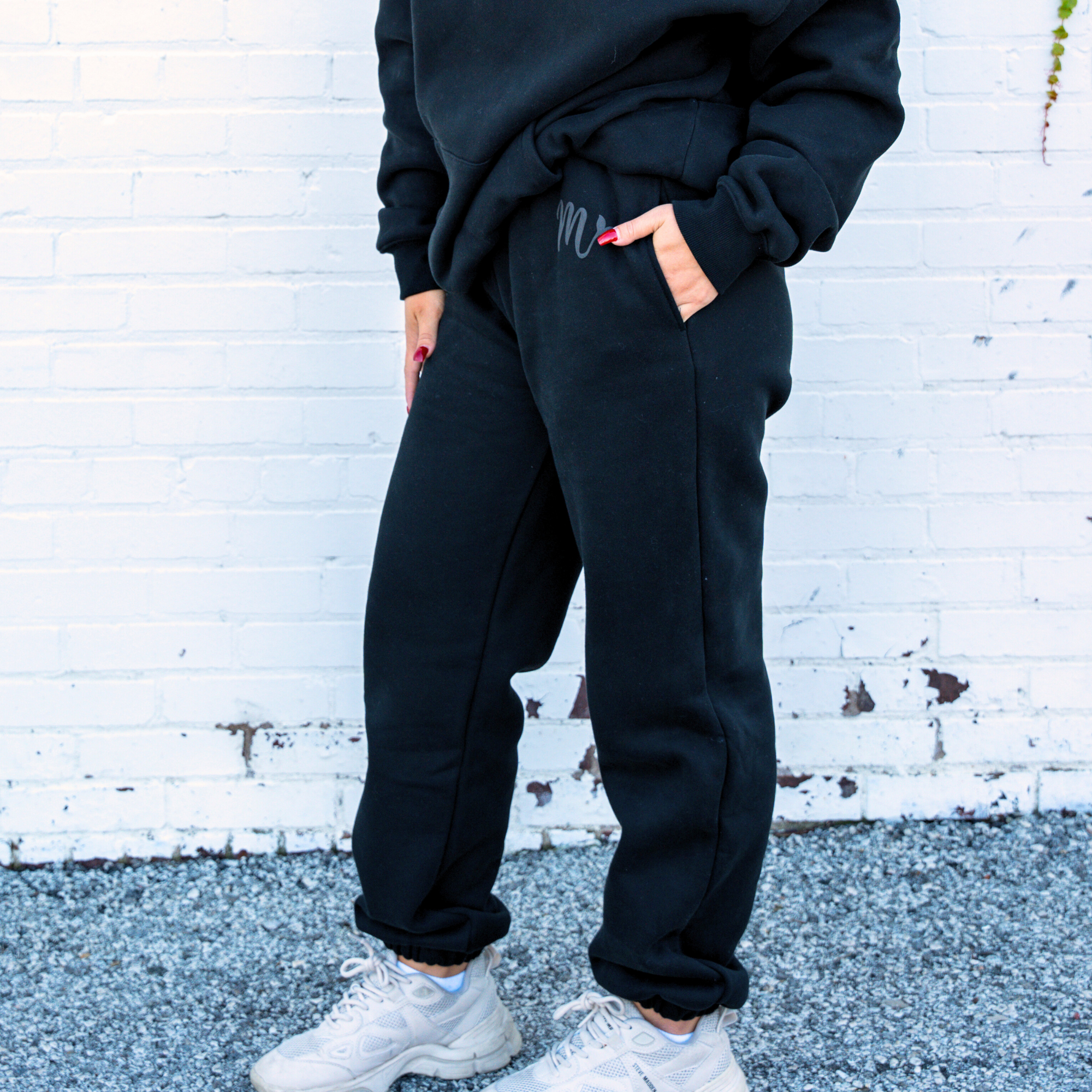 Model wearing black sweatpants paired with a matching black hoodie, both featuring a subtle embossed logo. The sweatpants have a relaxed fit with cuffed ankles and an elastic waistband, styled casually with white sneakers. The outfit exudes comfort and a sleek, minimalist look against a white brick background.